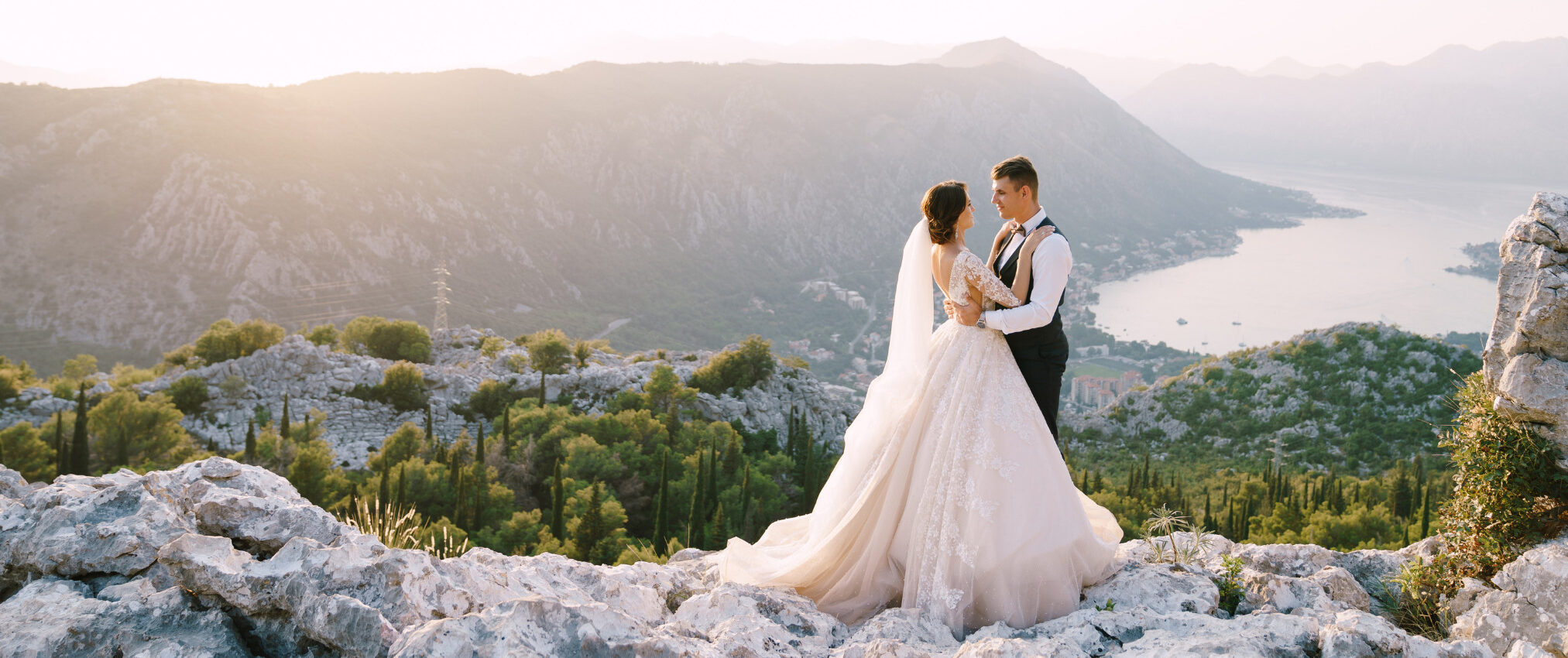 Bride and groom in formal attire standing on rocky cliff with spectacular view of mountains and lake below